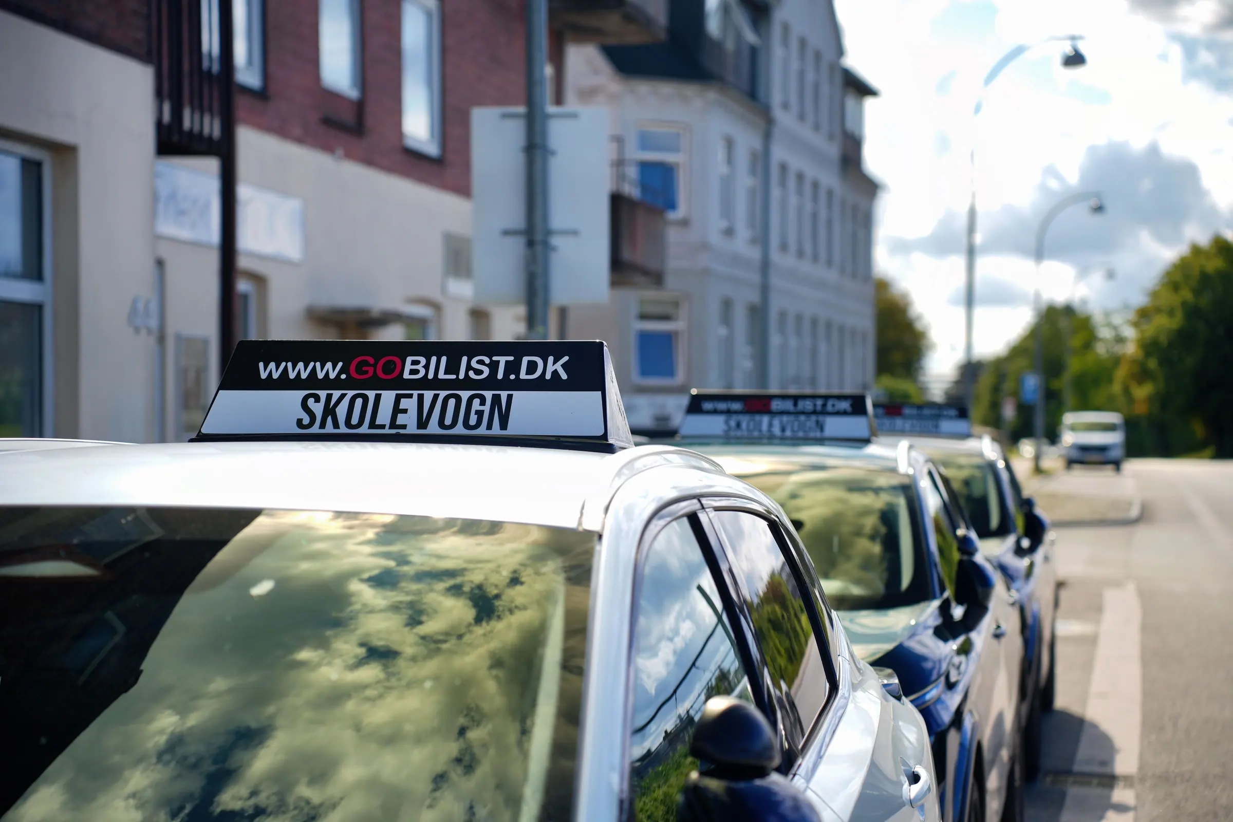 Tagskilt med GOBILIST.DK og teksten skolevogn på en hvid skolevogn, med flere skolevogne parkeret langs gaden